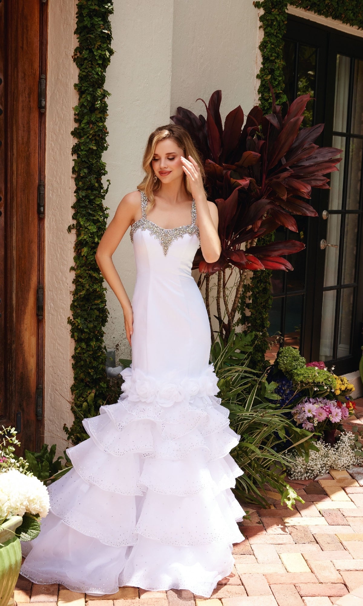 A woman wearing the Ava Presley 39566 Ruffle Mermaid Prom Dress, featuring beaded straps and a layered ruffle skirt, stands on a brick patio in front of a house with greenery and potted plants.